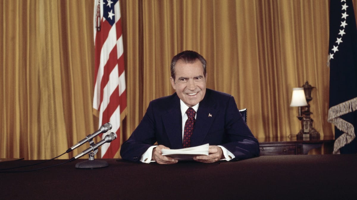 Richard Nixon smiling at his desk holding a stack of papers.